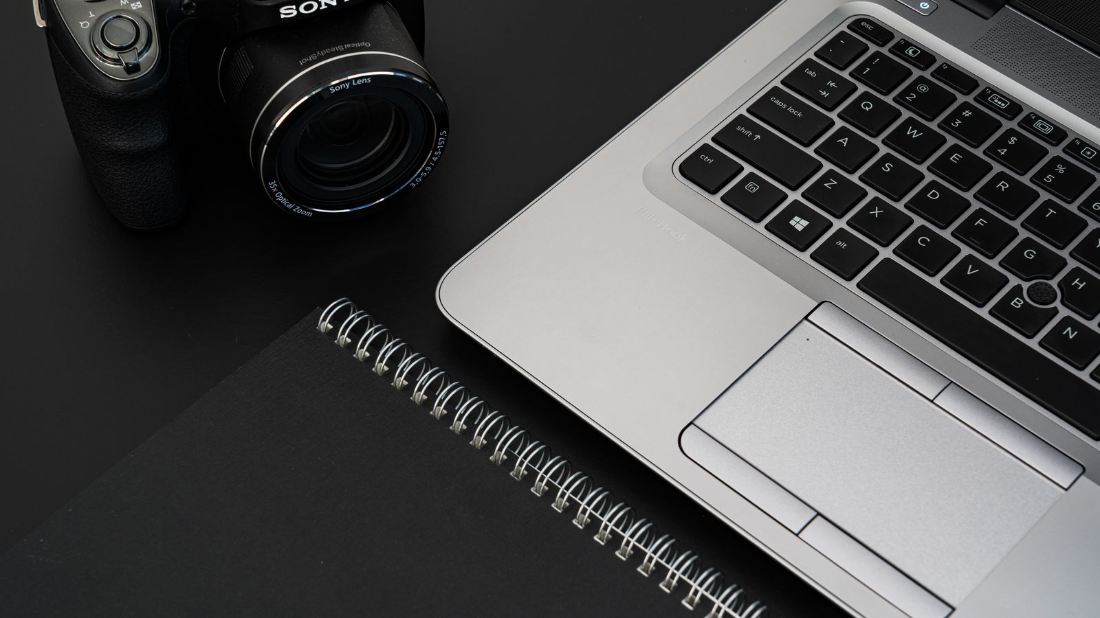A camera, laptop, and notebook on a dark desk.